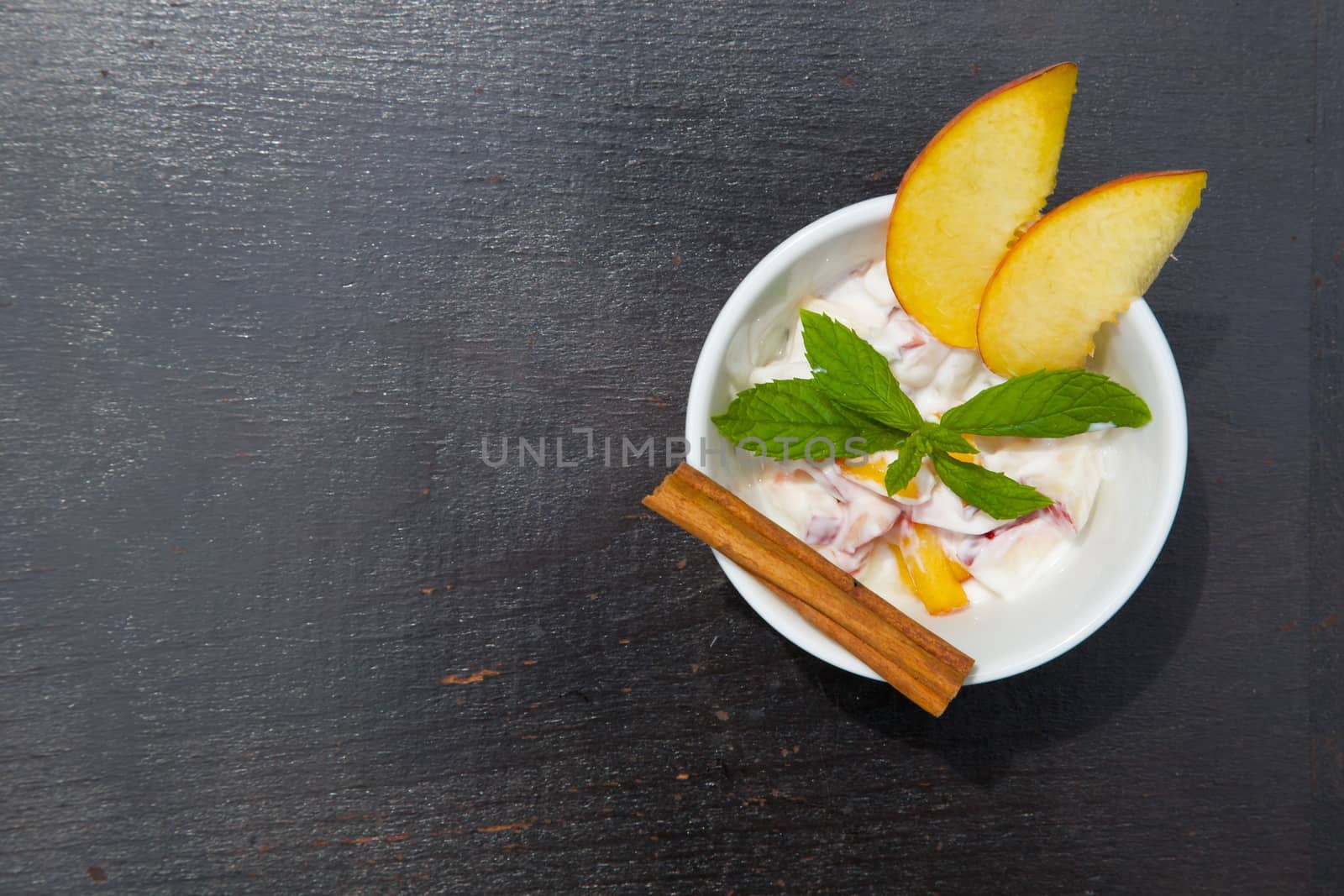 Homemade peach yogurt in a white dish on the black wooden surface. Decorated with two slices of fresh peach, fresh peppermint leaves and cinnamon sticks. Top view. Free space for a text