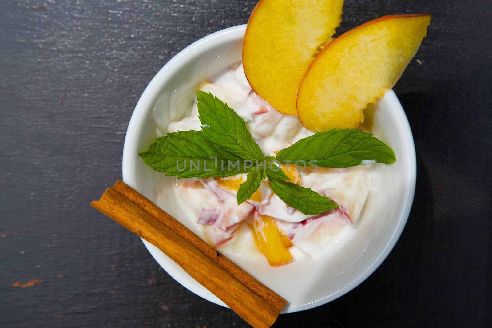 Homemade peach yogurt in a white dish on the black wooden surface. Decorated with two slices of fresh peach and fresh peppermint leaves and cinnamon stick. Top view. 