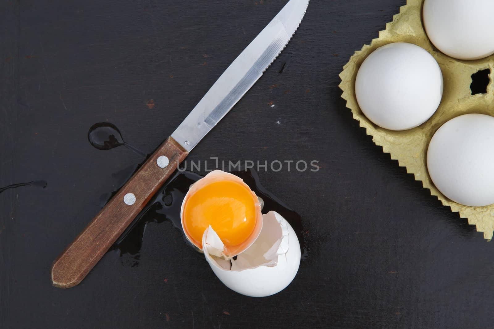 Broken egg, a knife with wooden handle and three white chicken eggs on the black wooden surface