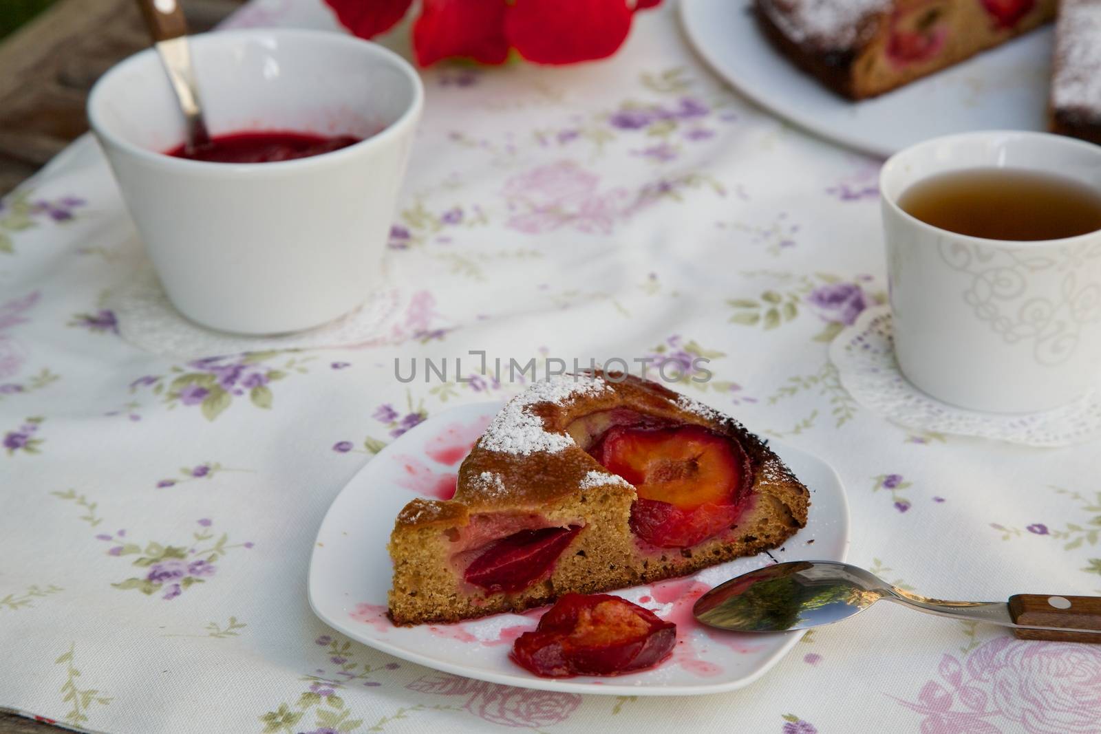 A piece of plum pie on a plate on a festive table. Plum pie, plum marmalade in a white dish and a cup of tea in the background