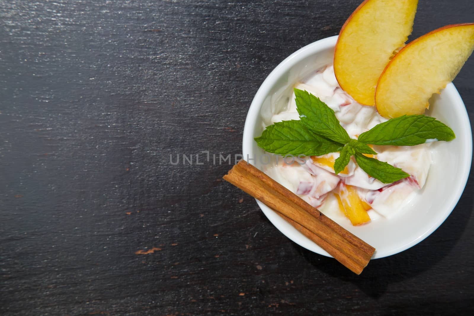 Homemade peach yogurt in a white dish on the black wooden surface. Decorated with two slices of fresh peach and fresh peppermint leaves and cinnamon stick. Top view. Free space for a text