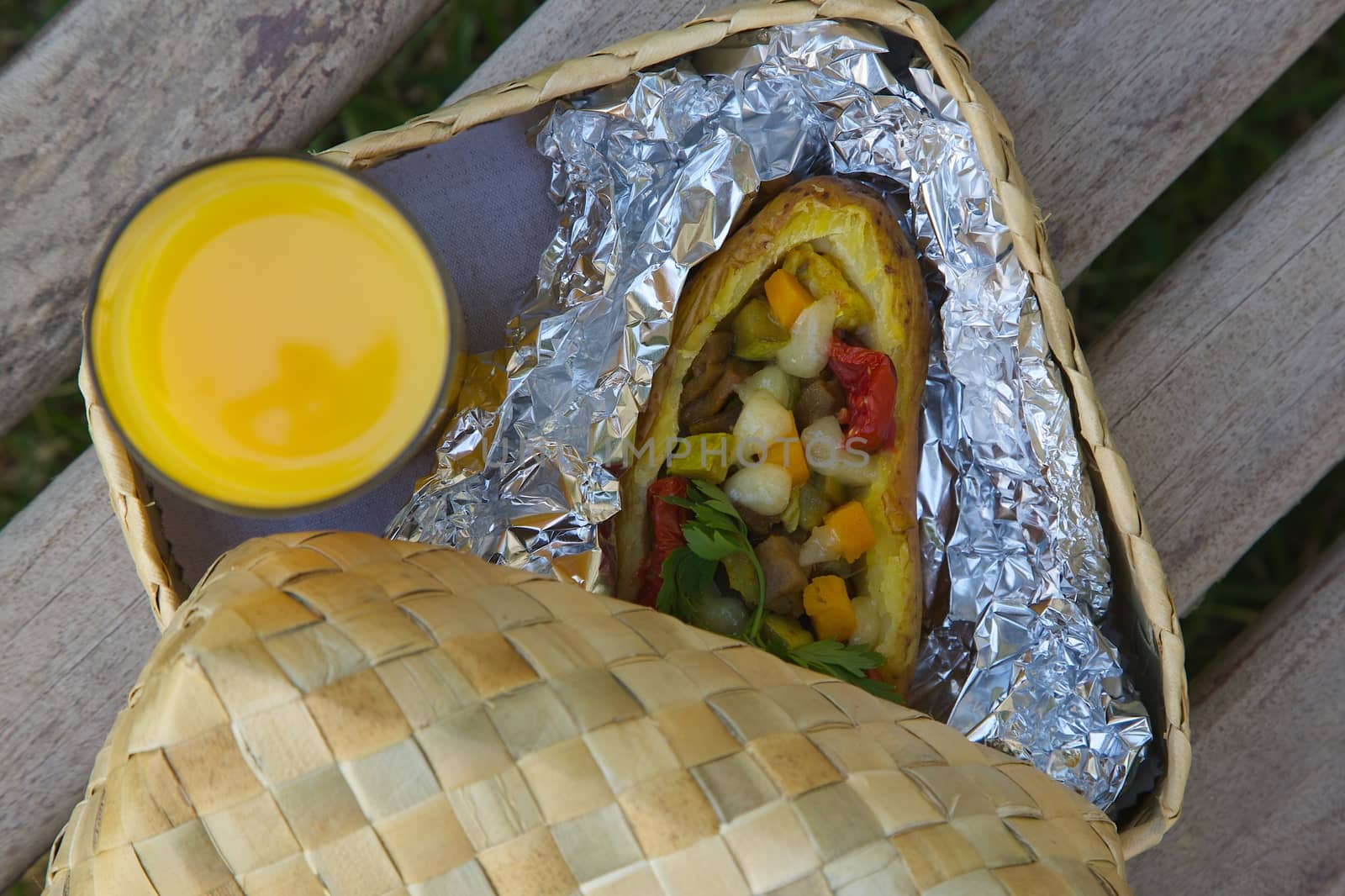 Open-air lunch for a vegetarian- potato baked with vegetables and cheese in an aluminium foil and a glass of fresh orange juice. The lunch is hidden in the woven birch basket