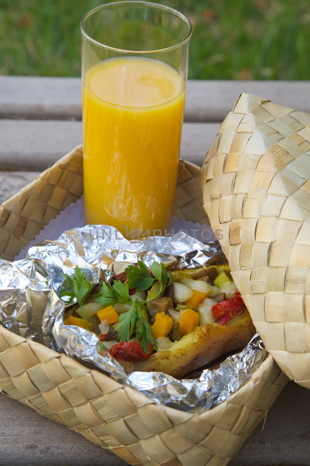 Open-air lunch for a vegetarian- potato baked with vegetables and cheese in an aluminium foil and a glass of fresh orange juice. Dish is hidden in the woven birch basket