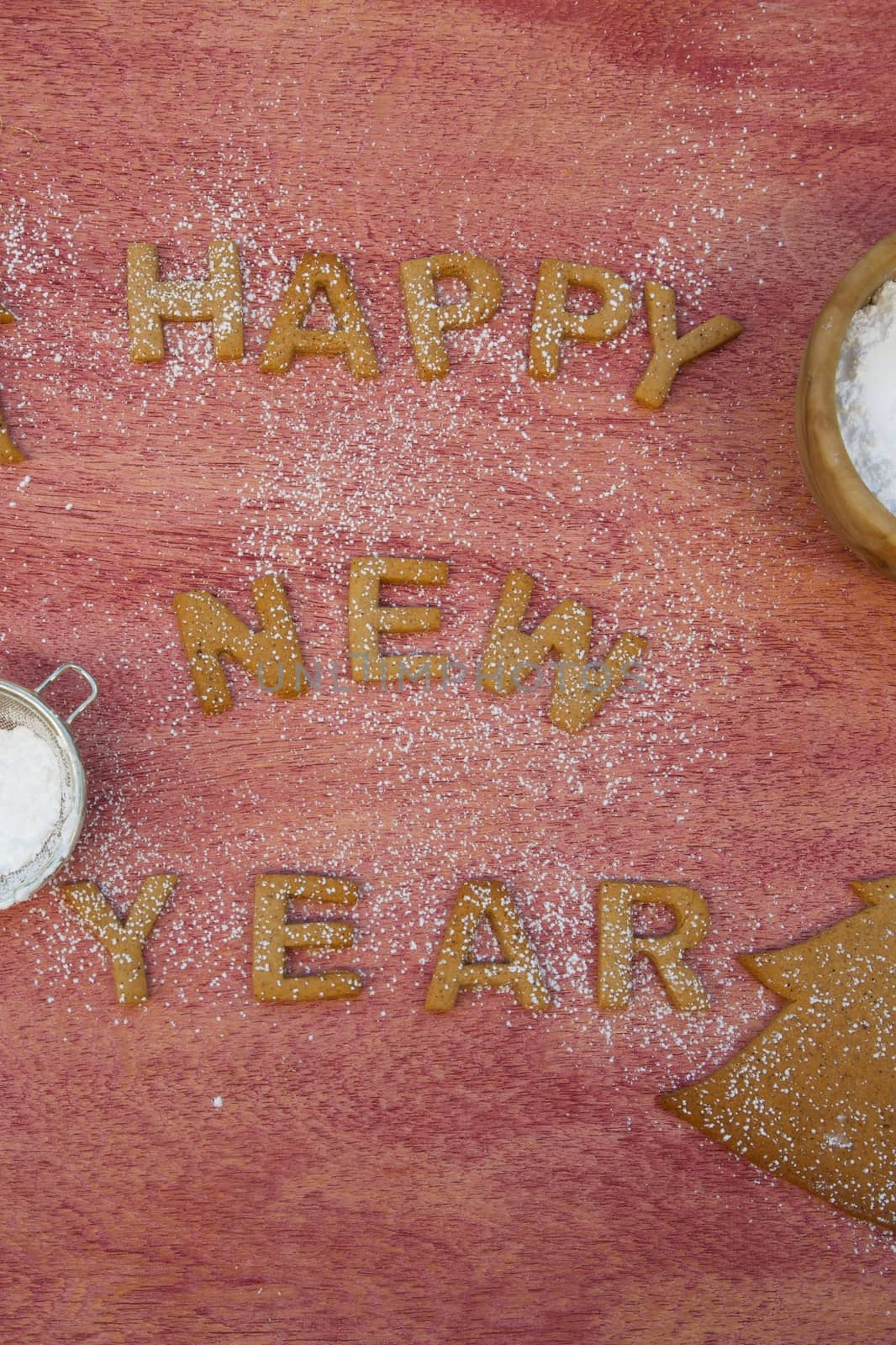 New Year's Day background with biscuit inscription: "Happy New Year". Tea strainer and olive wood dish with sugar powder on a red wooden surface
