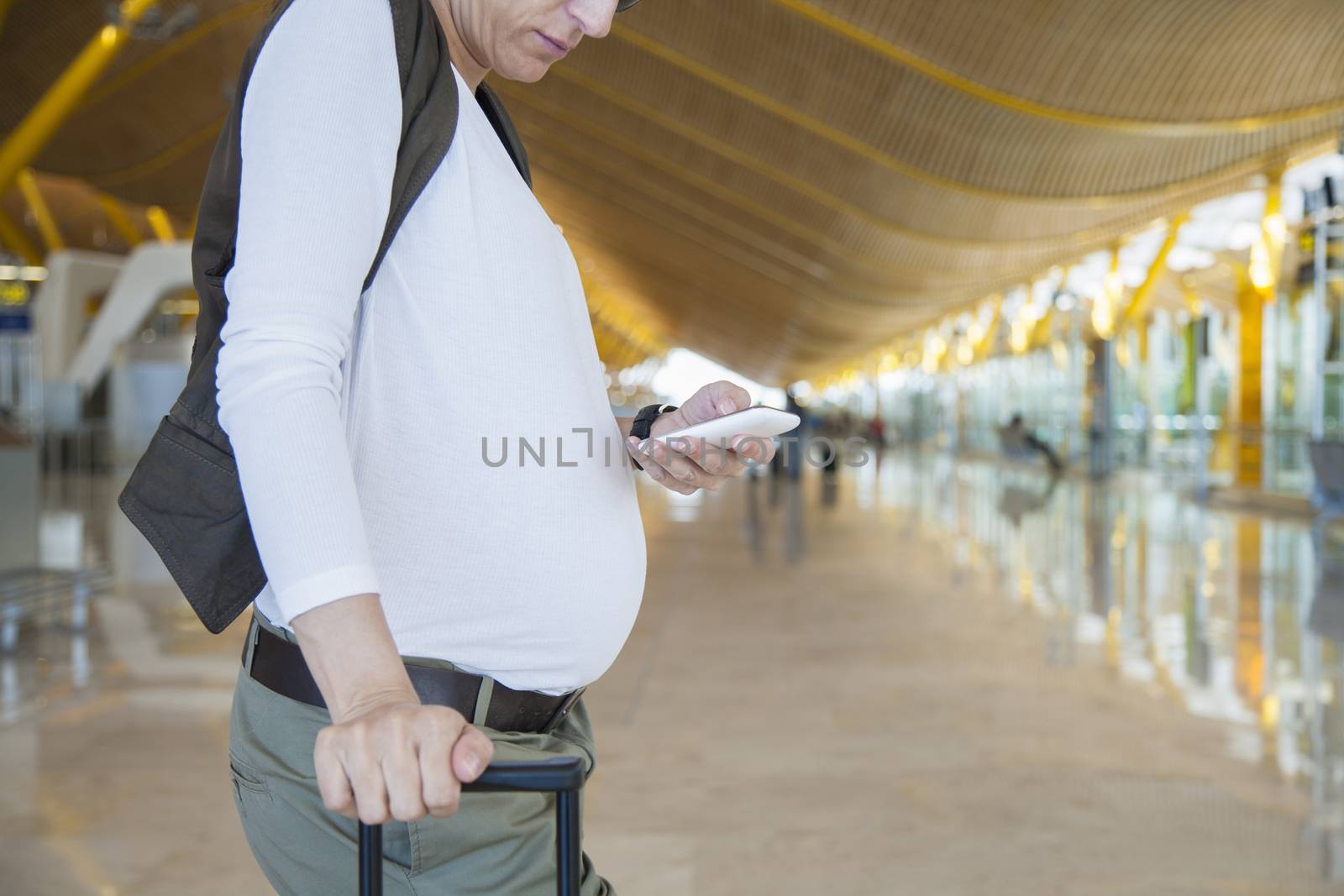 white shirt and vest woman pregnant touching phone in airport hall holding handle suitcase