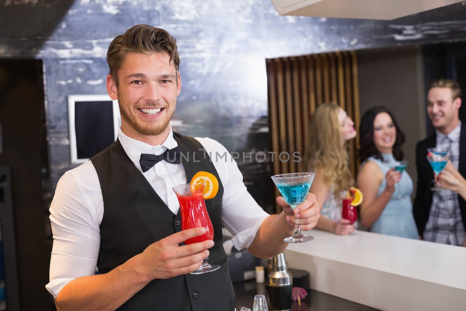 Handsome barman smiling at camera holding cocktails at the bar