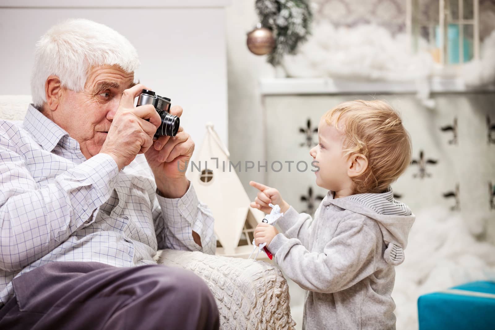 Senior man taking photo of his toddler grandson while sitting near Christmas tree at home