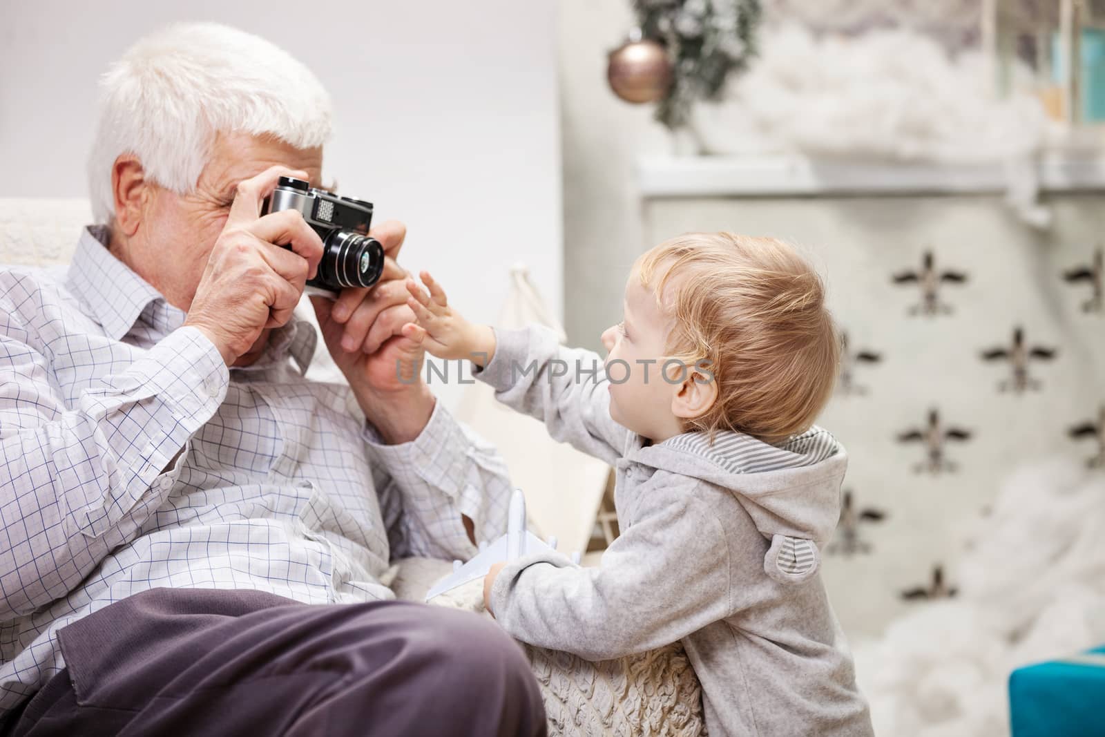 Senior man taking photo of his toddler grandson while sitting near Christmas tree at home