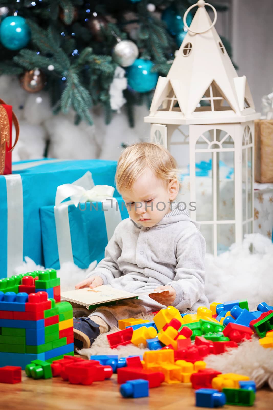 Cute toddler boy sitting at Christmas tree and reading book. Building blocks scattered around.