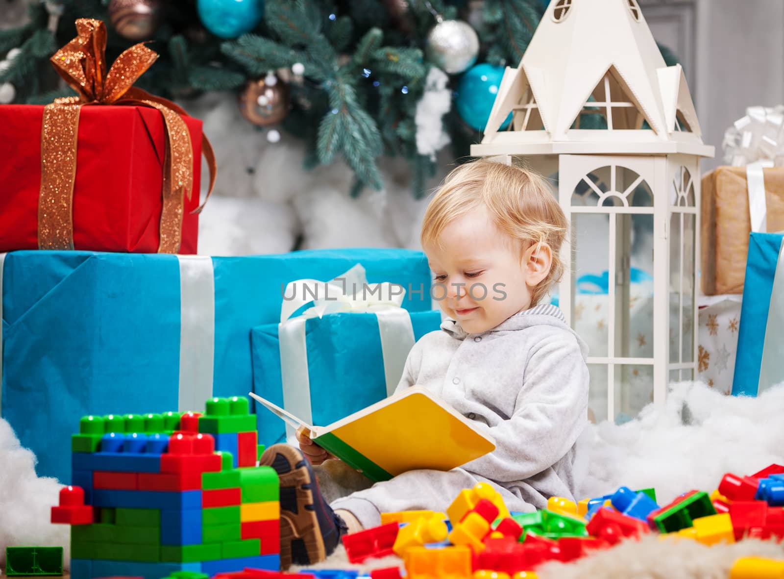 Cute toddler boy sitting at Christmas tree and reading book. Building blocks scattered around.