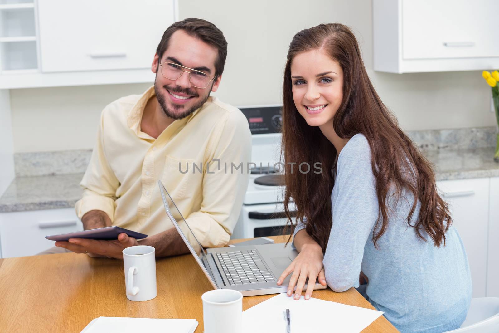 Young couple smiling at camera using technology at home in the kitchen