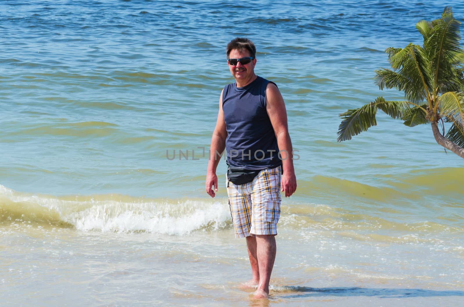 Man in shorts, T-shirt and sunglasses on the beach with your feet in the water.