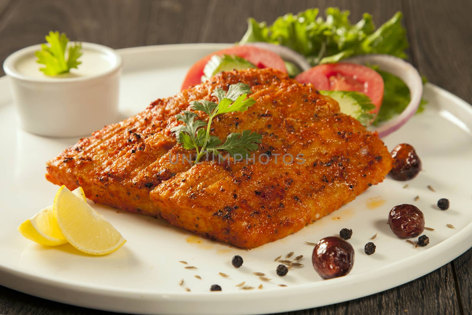 Grilled fish tikka served on a plate with salad and tarter sauce dip and salad with lemon, on a dark wood background