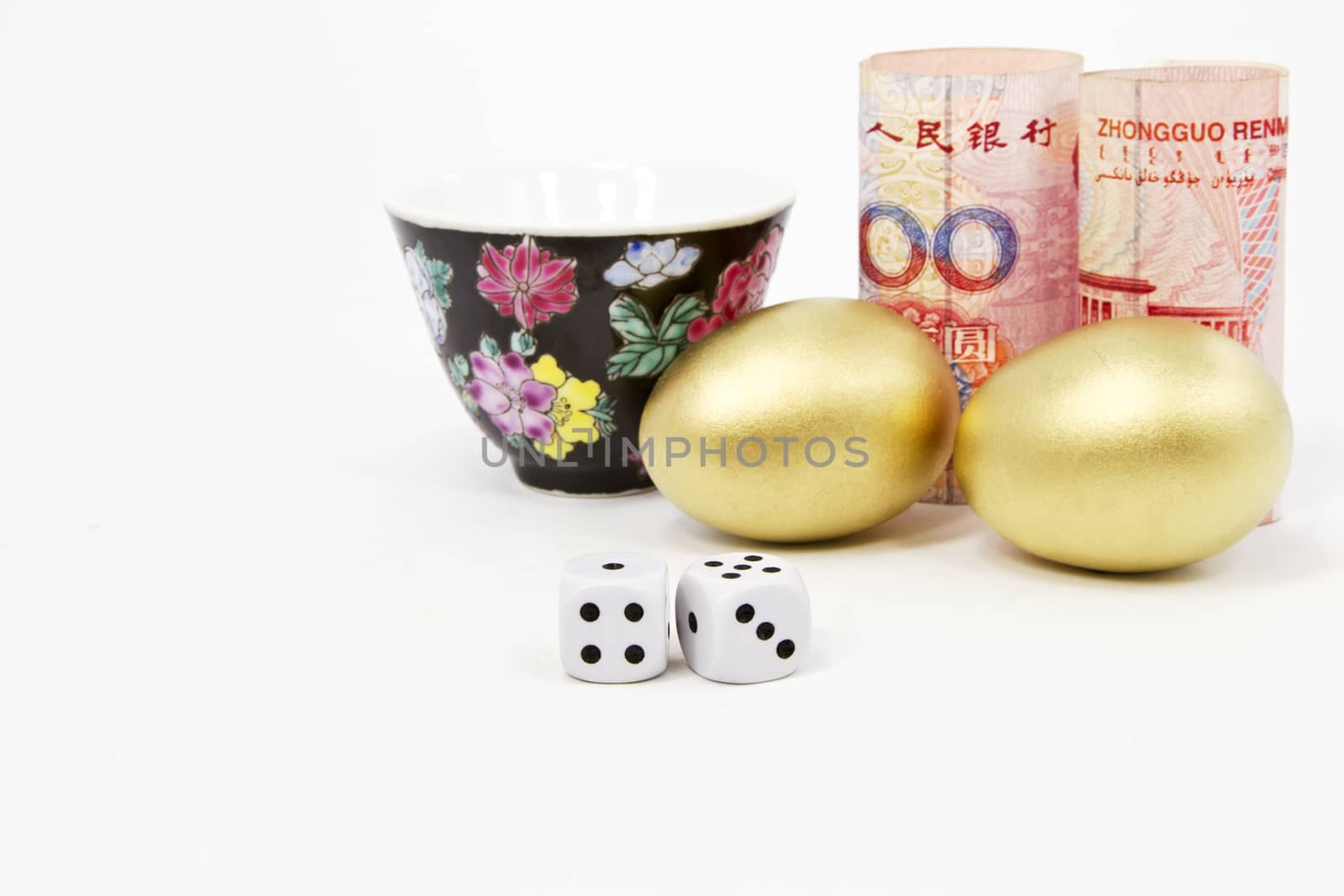 Dice sit in front of Chinese yuan currency, two, gold nest eggs, and porcelain tea mug in a financial investment concepts still life suggestive of Asian influences, perhaps risk, on the world economy and investment strategies.  