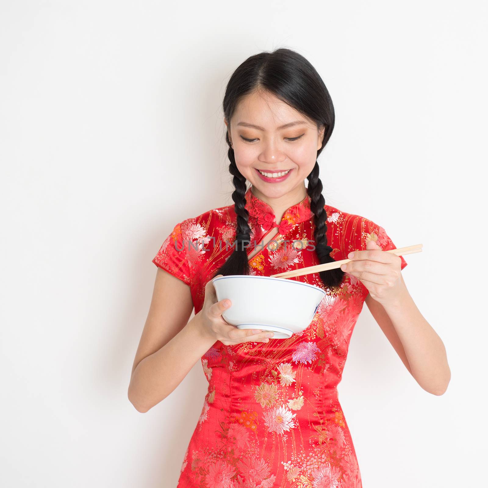 Portrait of Asian Chinese female eating, using chopsticks holding rice bowl, in traditional dress red qipao standing on plain background.