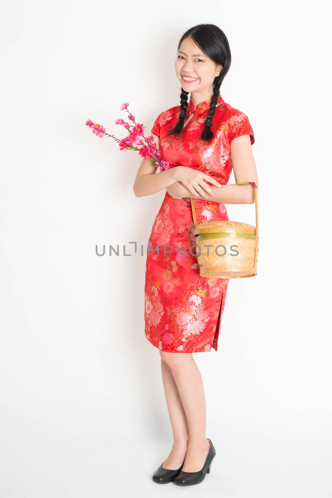Portrait of full length Asian Chinese girl hands holding gift basket and plum blossom, in traditional red qipao standing on plain background.