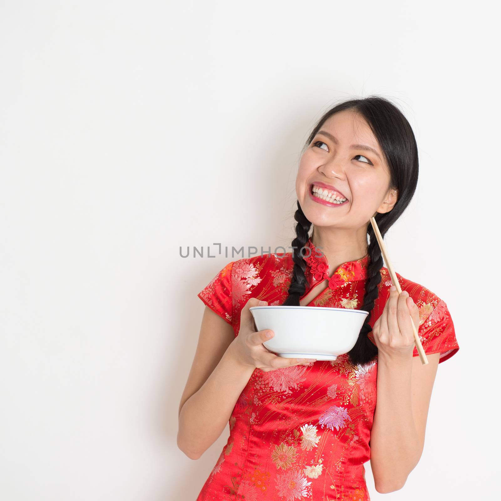 Asian Chinese female eating using chopsticks holding rice bowl, in traditional qipao on plain background, thinking and looking up at copy space.