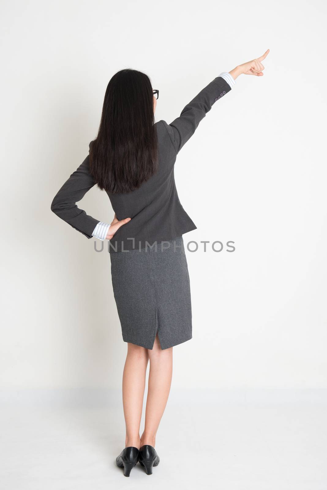 Full body rear view Asian business woman pointing at copy space, standing on plain background.