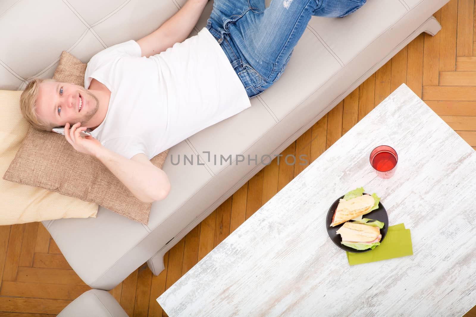 Young man with a Sandwich on the Sofa while talking on the phone.
