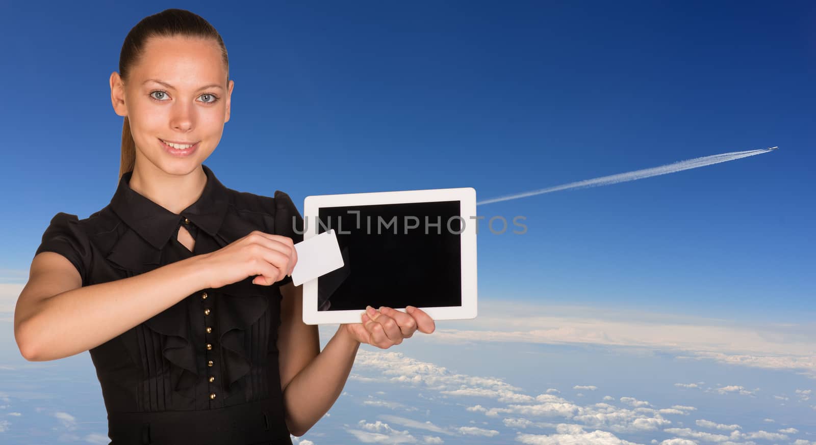 Beautiful businesswoman holding blank tablet PC and blank business card in front of PC screen.Blue sky, clowds and distant jet airliner with vapour trail as backdrop