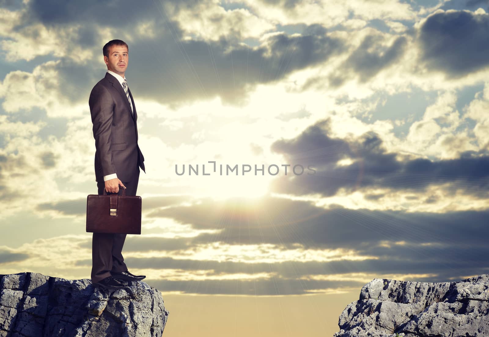 Businesswoman standing on the edge of rock gap, looking at camera as if uncertain. Sky with sun shining through clouds as backdrop