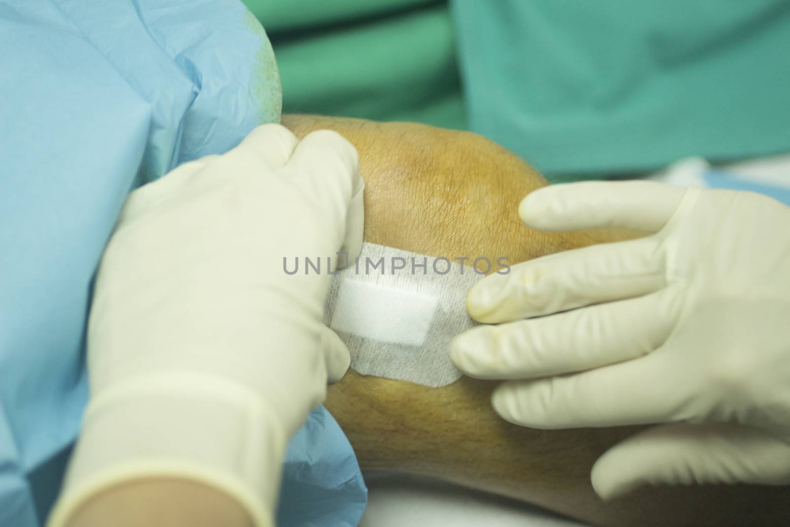 Close-up color photo of a female hospital clinic nurse wearing sterile blue gloves and senior male patient aged 65-70 being cleaned after receiving a an injection of human growth factors in his knee with sterilising liquid covering skin and sterile green hospital bed defocused background.