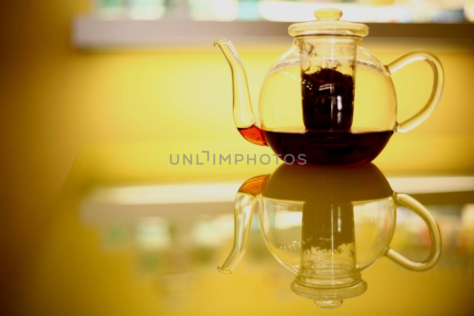 Teapot of transparent glass on glass kitchen table with herbal tea remedy with reflection in yellow tones. 