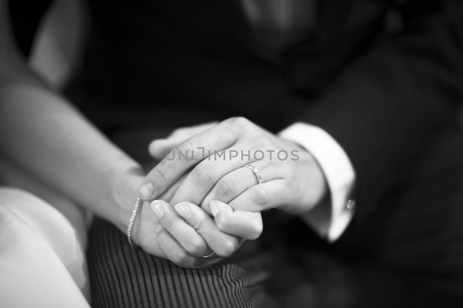 Black and white artistic digital photo of bridegroom in dark suit and white shirt in church religious wedding marriage ceremony holding hands with the bride in white long wedding bridal dress. Shallow depth of with background out of focus. 