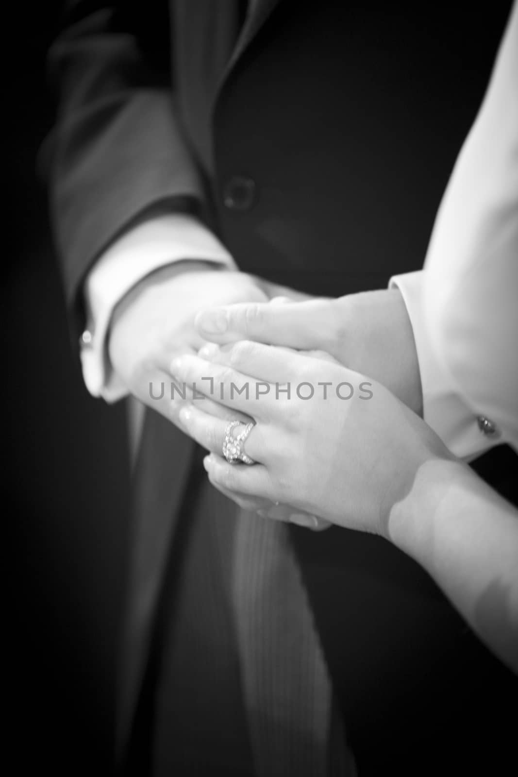 Black and white artistic digital photo of bridegroom in dark suit and white shirt in church religious wedding marriage ceremony holding hands with the bride in white long wedding bridal dress. Shallow depth of with background out of focus. 