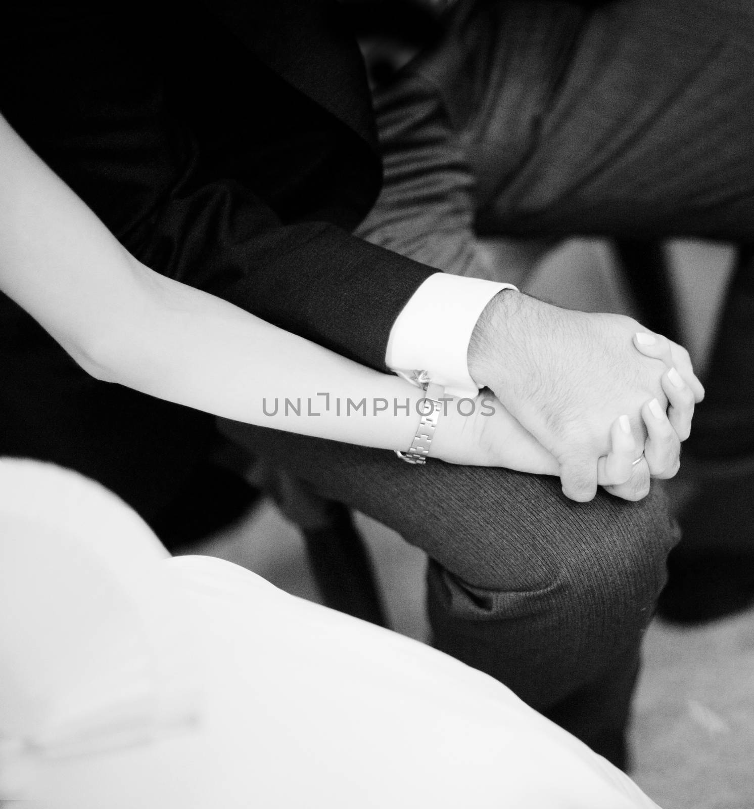 Black and white artistic digital rectangular horizontal photo of the bride wearing white wedding dress and bridegroom sitting holding hands during church wedding marriage ceremony in Madrid Spain. Shallow depth of on foreground with background out of focus.