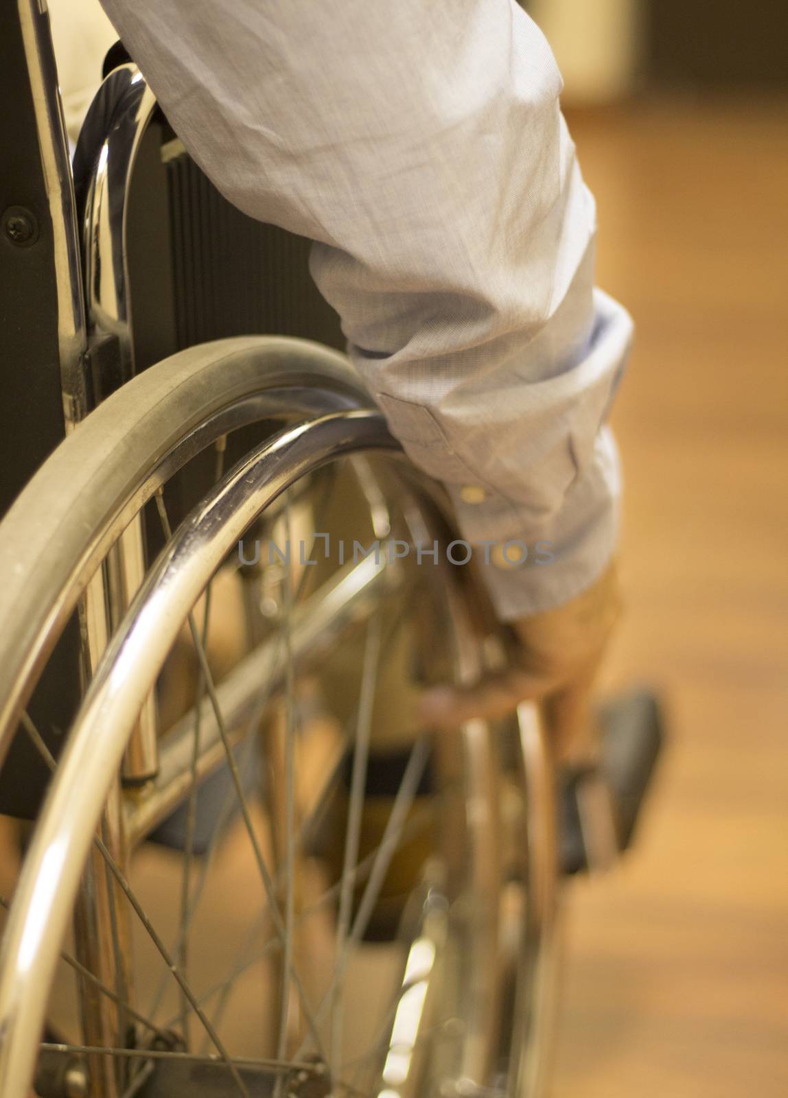 Man sitting in wheel chair wearing blue shirt pushing wheel to move himself forward in hospital clinic. Color digital photo in warm tones shot from behind with shallow depth of focus.