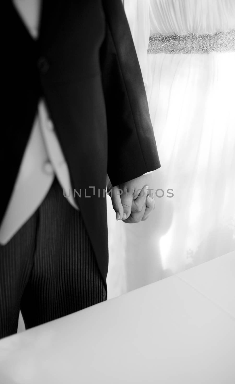 Black and white artistic digital rectangular horizontal photo of the bride wearing white wedding dress and bridegroom standing wearing long coat morning suit with waste coat holding hands during church wedding marriage ceremony in Madrid Spain. Shallow depth of on foreground with background out of focus. 