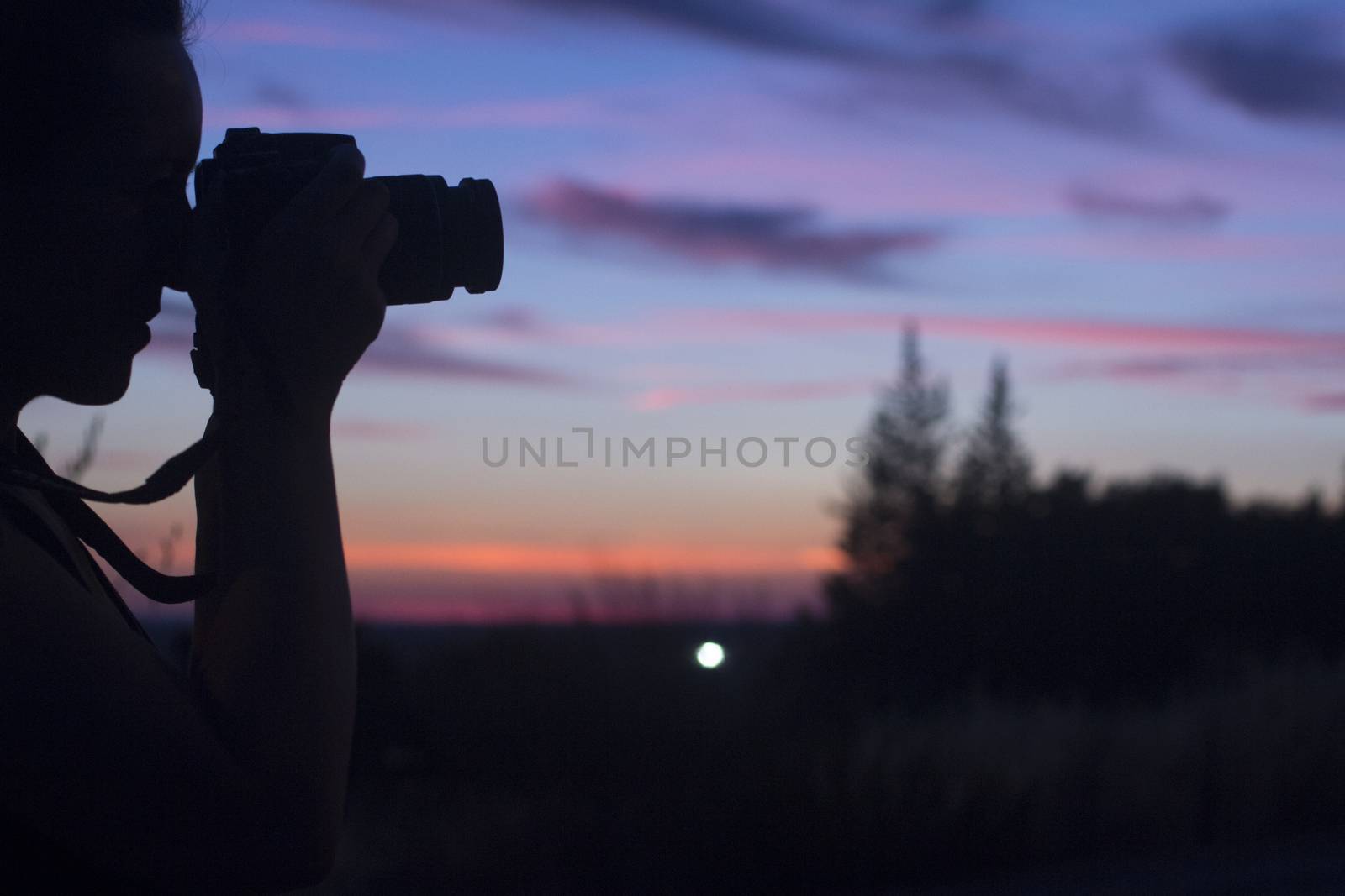 Female photographer taking photo in silhouette against blue purple sky with medium format analogue film camera and strap with long lens and filter holder on hot summer evening. 