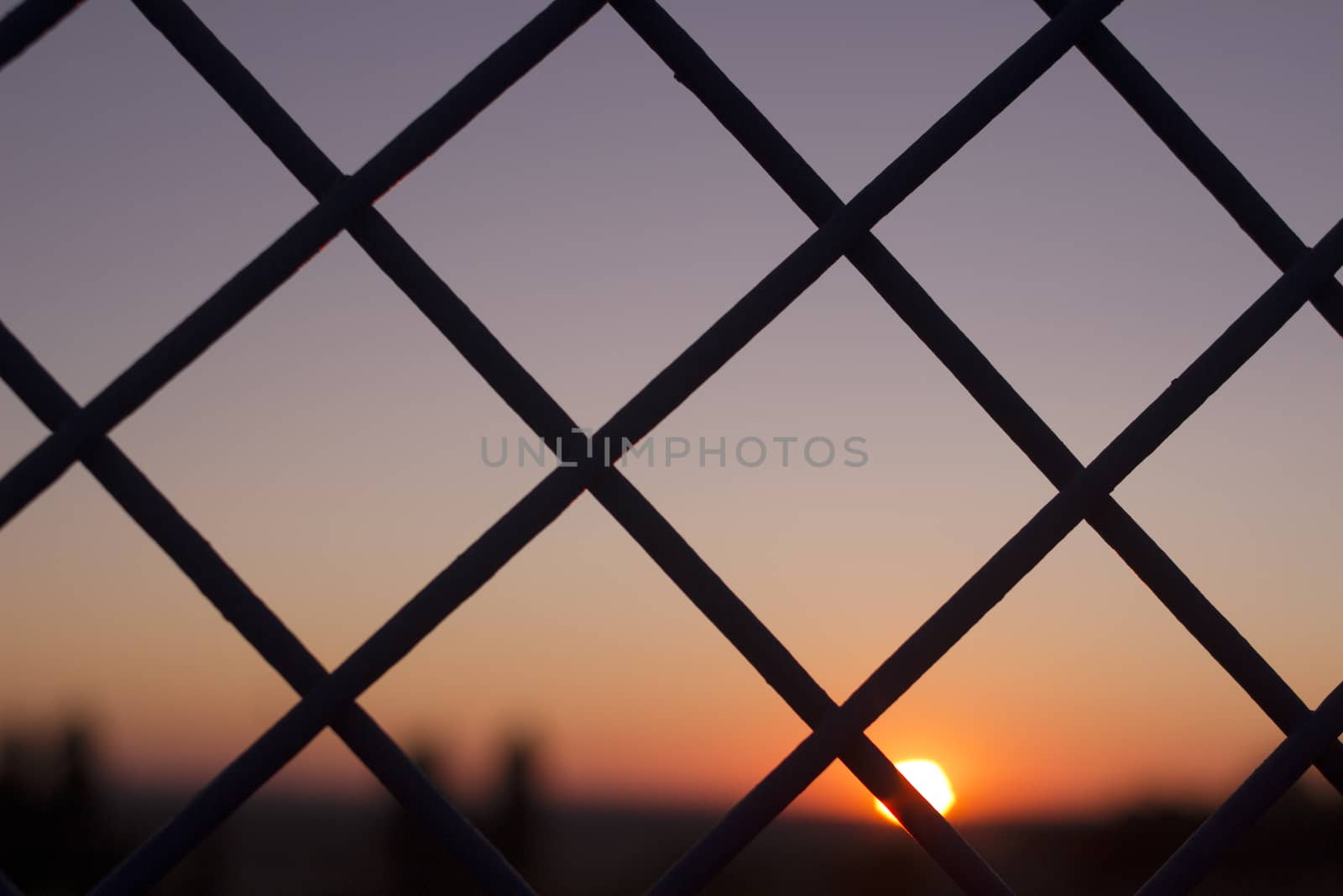 Horizontal color digital photo of setting evening sun and red and blue sky shining through a metal wire mesh fence in silhouette at dusk in dark tones with bokeh blur shallow depth of focus defocused landscape background. 