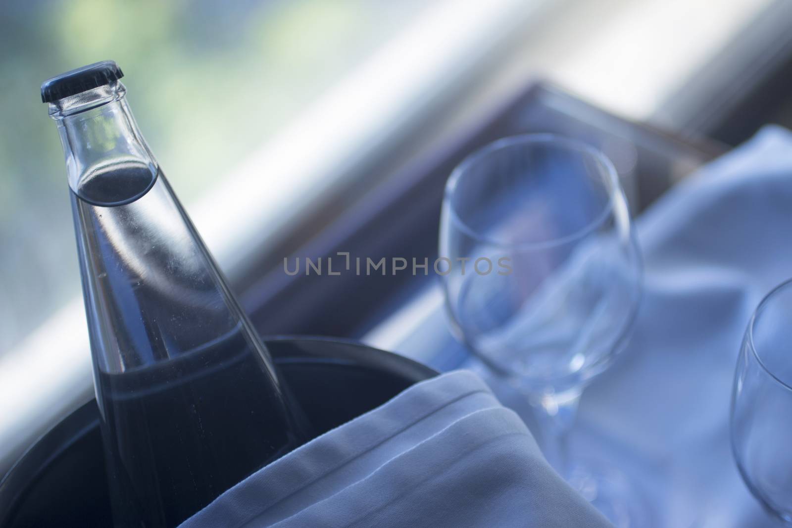 Artistic still life photo of two glasses and bottle of water in metal ice bucket with cloth covering on a glass table in room with window behind in soft evening purple blue color light with out of focus in background.