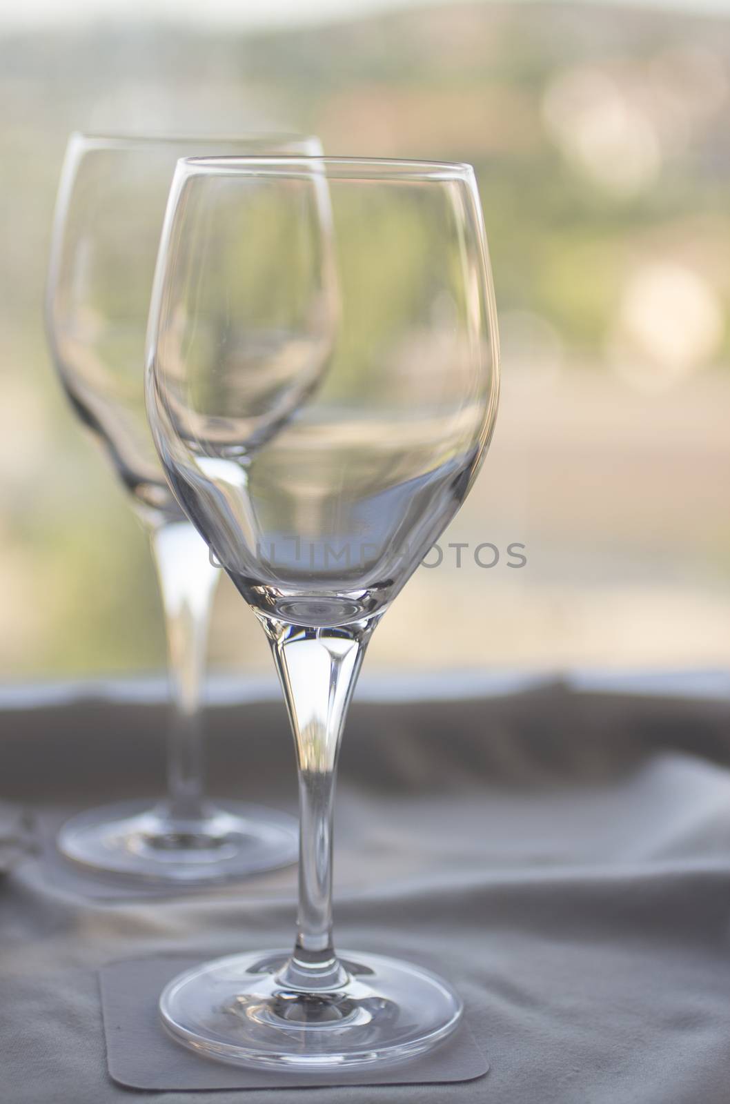 Still life photo of two wine or water glasses on coasters on cloth on tray on a table in room with window behind in soft evening pastel color light out of focus in background. 