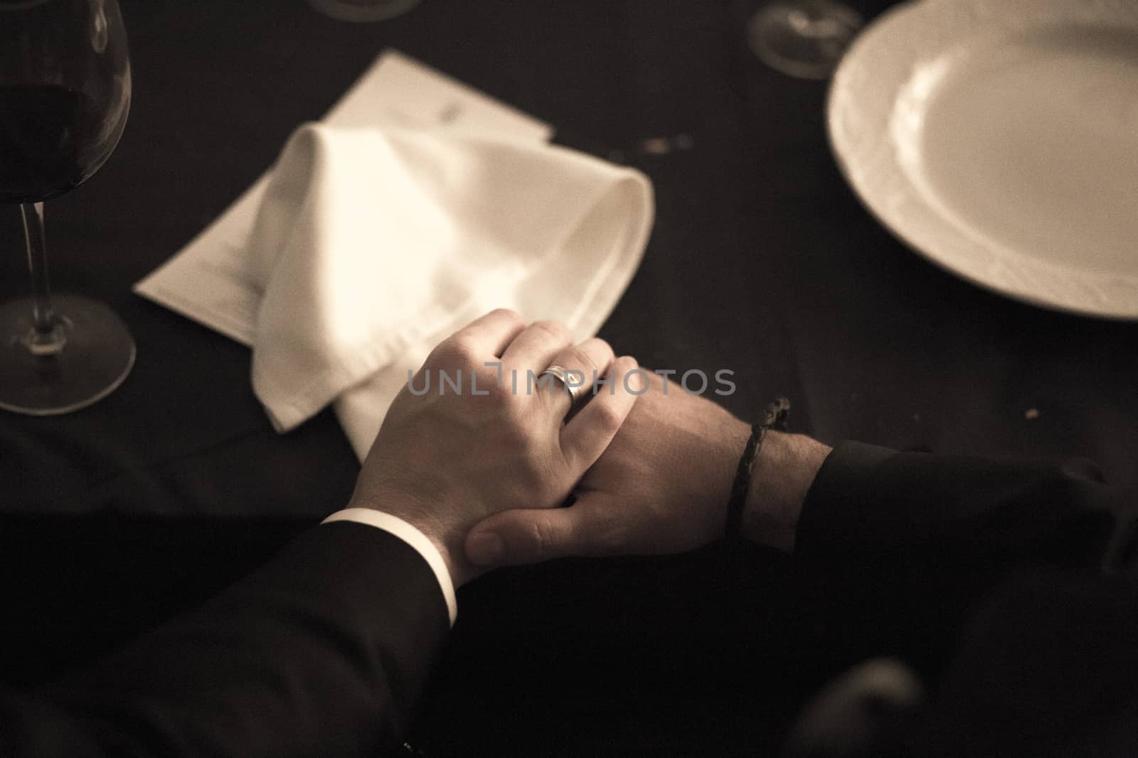 Color artistic digital rectangular horizontal photo of the bride and groom holding hands on the white tablecloth of the banquet dinner table with plates and glasses in a wedding party in a luxury hotel in Spain. Shallow depth of with background out of focus.