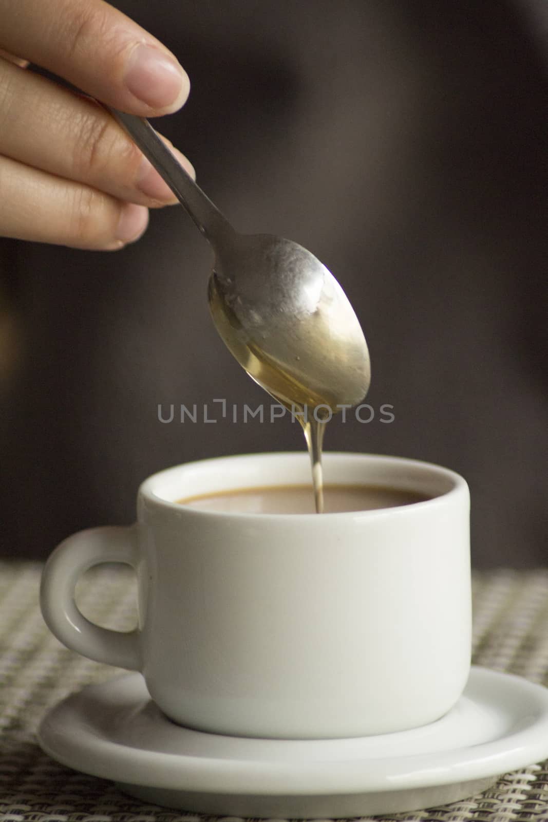 Hand of young woman ading corn syrup with spoon in cup of coffee expresso with milk on saucer in table.