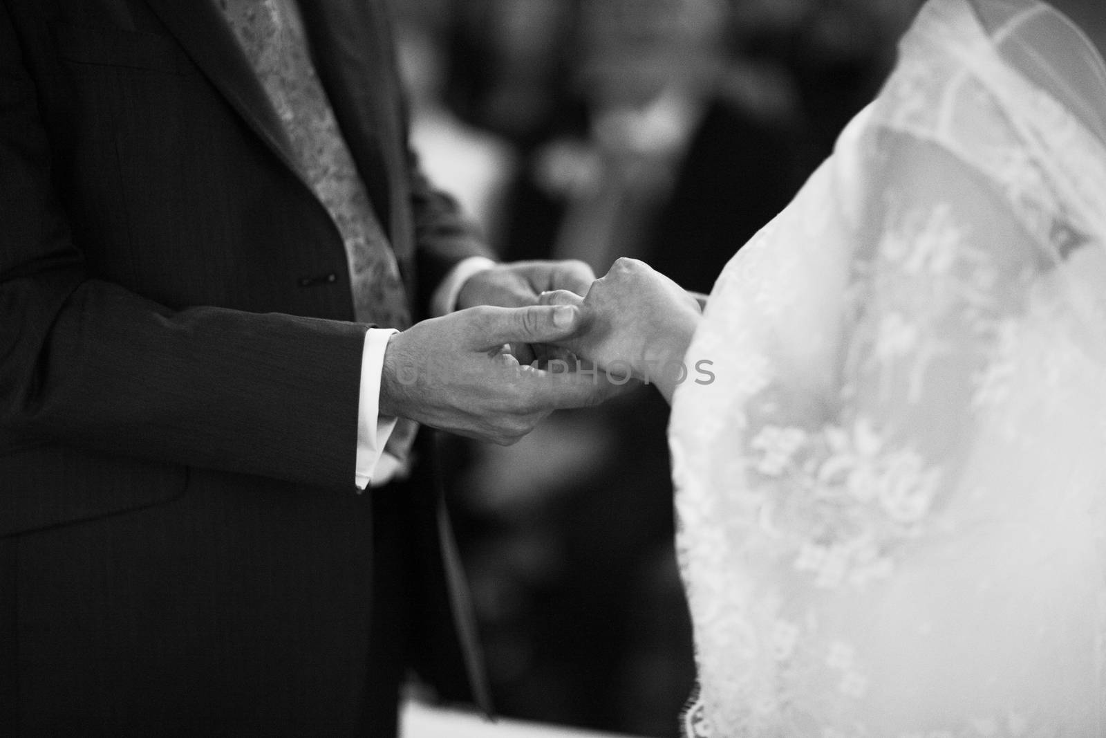 Black and white artistic digital rectangular horizontal photo of hand of bridegroom in dark long morning suit and white shirt with cufflinks in church religious wedding marriage ceremony holding hands to exchange wedding rings with the bride in white long wedding bridal dress in Barcelona Spain. Shallow depth of with background out of focus. 
