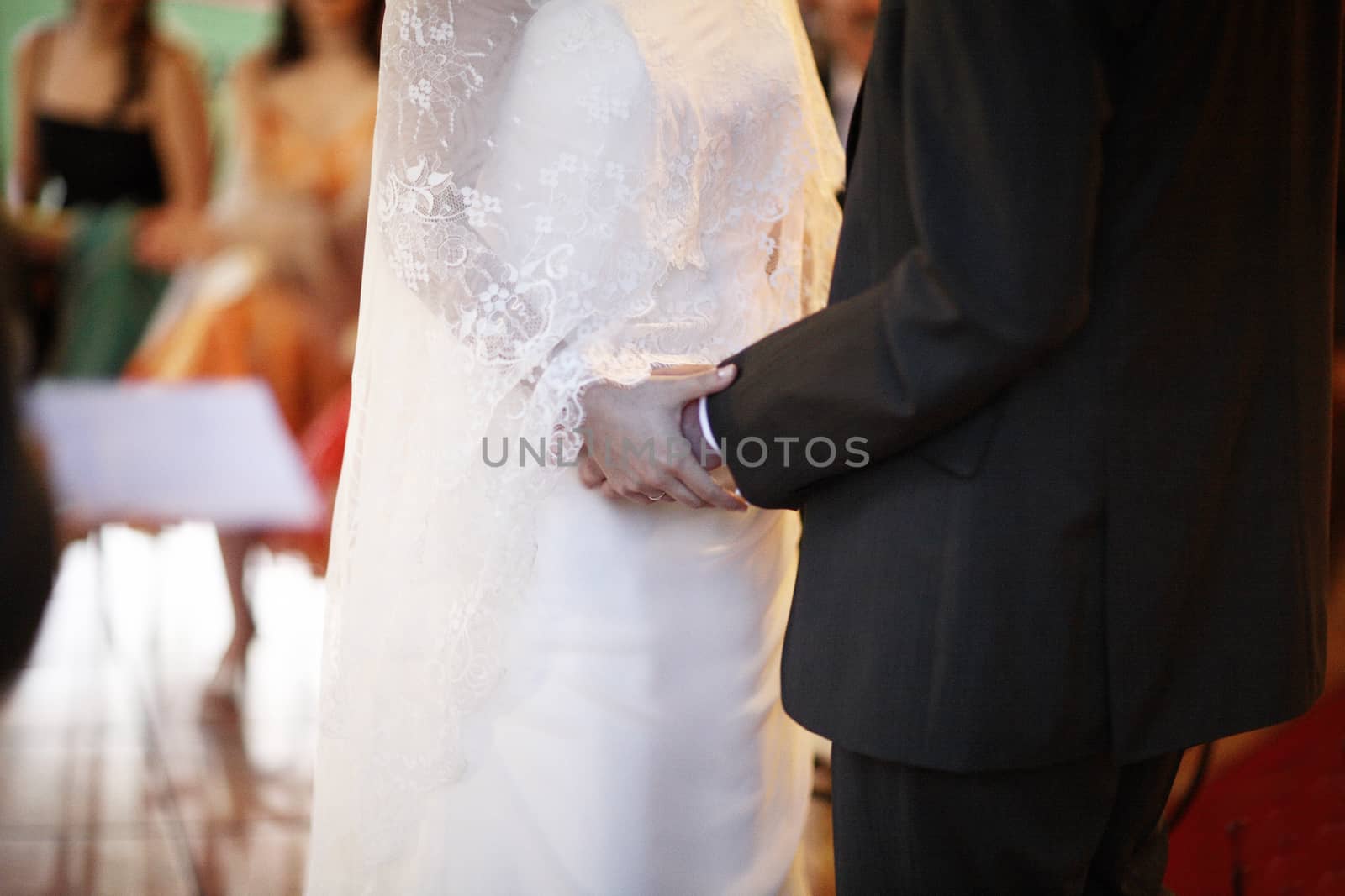 Color artistic digital photo of bridegroom in dark suit and white shirt with cufflinks in church religious wedding marriage ceremony holding hands with the bride in white wedding bridal dress. Shallow depth of with background out of focus. 