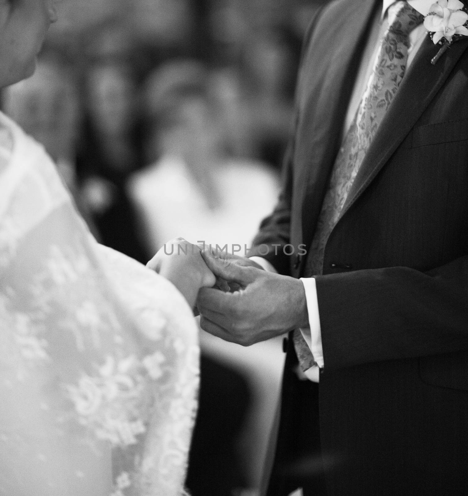 Black and white artistic digital rectangular horizontal photo of hand of bridegroom in dark long morning suit and white shirt with cufflinks in church religious wedding marriage ceremony holding hands to exchange wedding rings with the bride in white long wedding bridal dress in Barcelona Spain. Shallow depth of with background out of focus. 