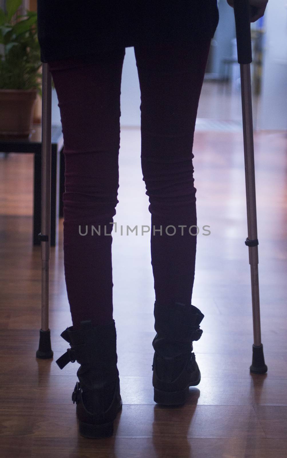 Young woman in black jeans and boots walking on aluminium metal crutches in hospital clinic in artistic silhouette color photo.