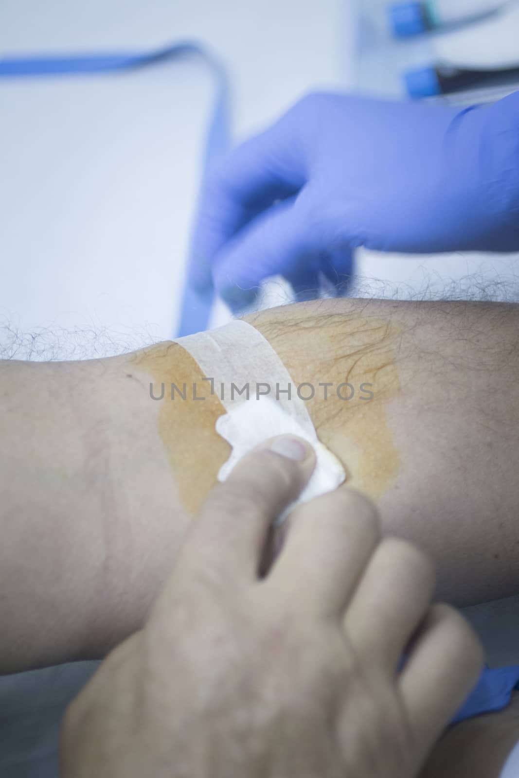 Close-up color photo of a female hospital clinic nurse wearing sterile blue gloves and senior male patient aged 65-70 giving a blood donation sample from the interior of his elbow resting arm on sterile blue white defocused background.