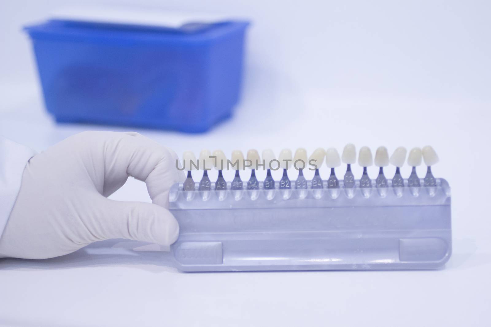 Hand of dental nurse in white sterile surgical glove holding the dentist's tooth color guide to choose appropriate natural colors for a patient's tooth implants and crowns on plain white pastel blue background of the dental surgery clinic table. 