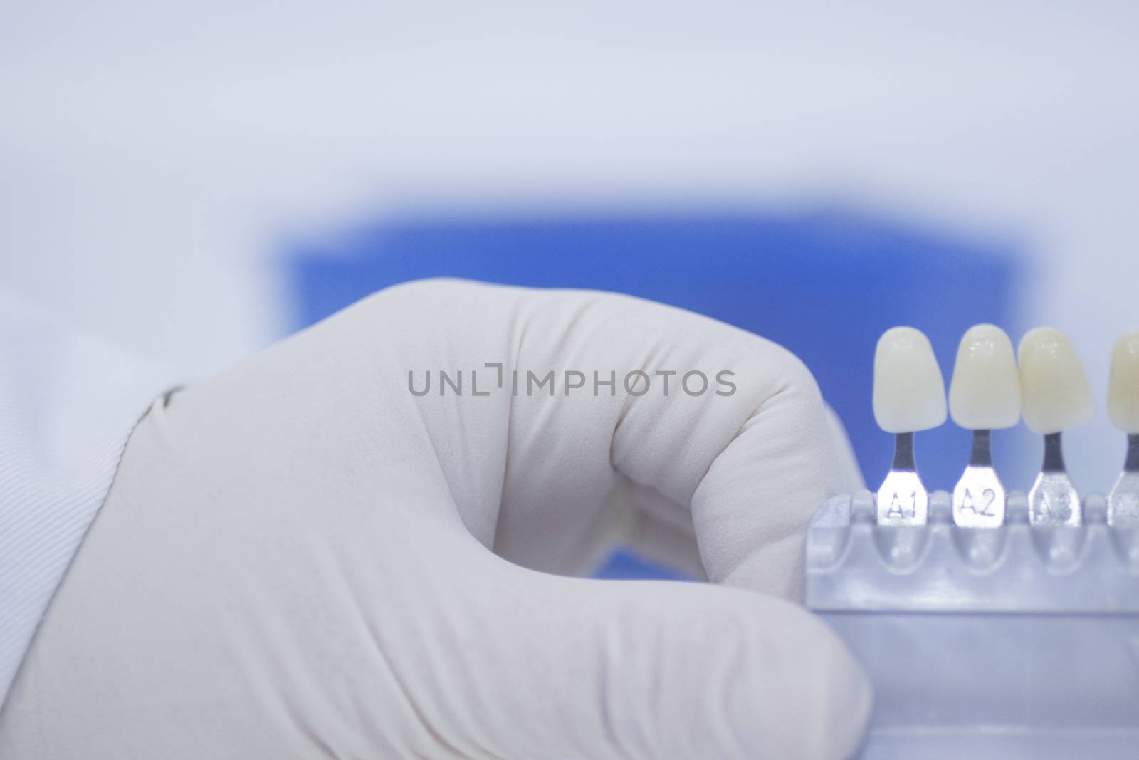 Hand of dental nurse in white sterile surgical glove holding the dentist's tooth color guide to choose appropriate natural colors for a patient's tooth implants and crowns on plain white pastel blue background of the dental surgery clinic table. 