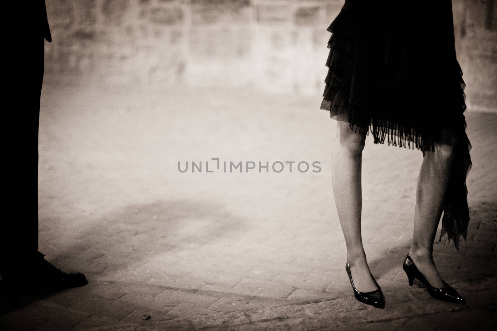 Legs of young lady wearing high heels shoes and man in suit standing outside in social event wedding marriage party in Madrid Spain at night. black and white sepia tone photograph. 