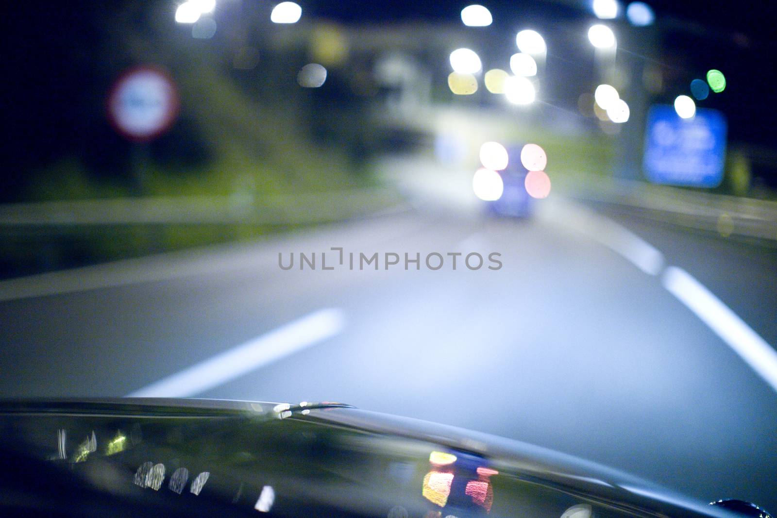 Road at night seen from inside car driving on motorway with highway and city lights in bokeh out of focus with shallow depth of focus on bonnet. 