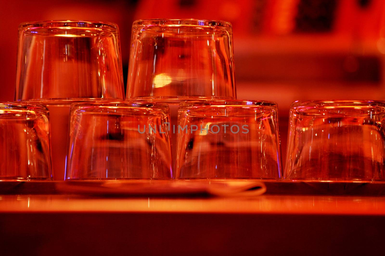 Color photo in red tones of a group of upturned clear wine glasses sparkling in the light in a pub / public house restaurant bar at night in Chueca in Madrid Spain. The multiple glasses have just been washed and dried, and are displayed stacked upside-down on the top of the bar where they are reflecting the light from above, ready to be filled with wine and served to the evening's customers. 