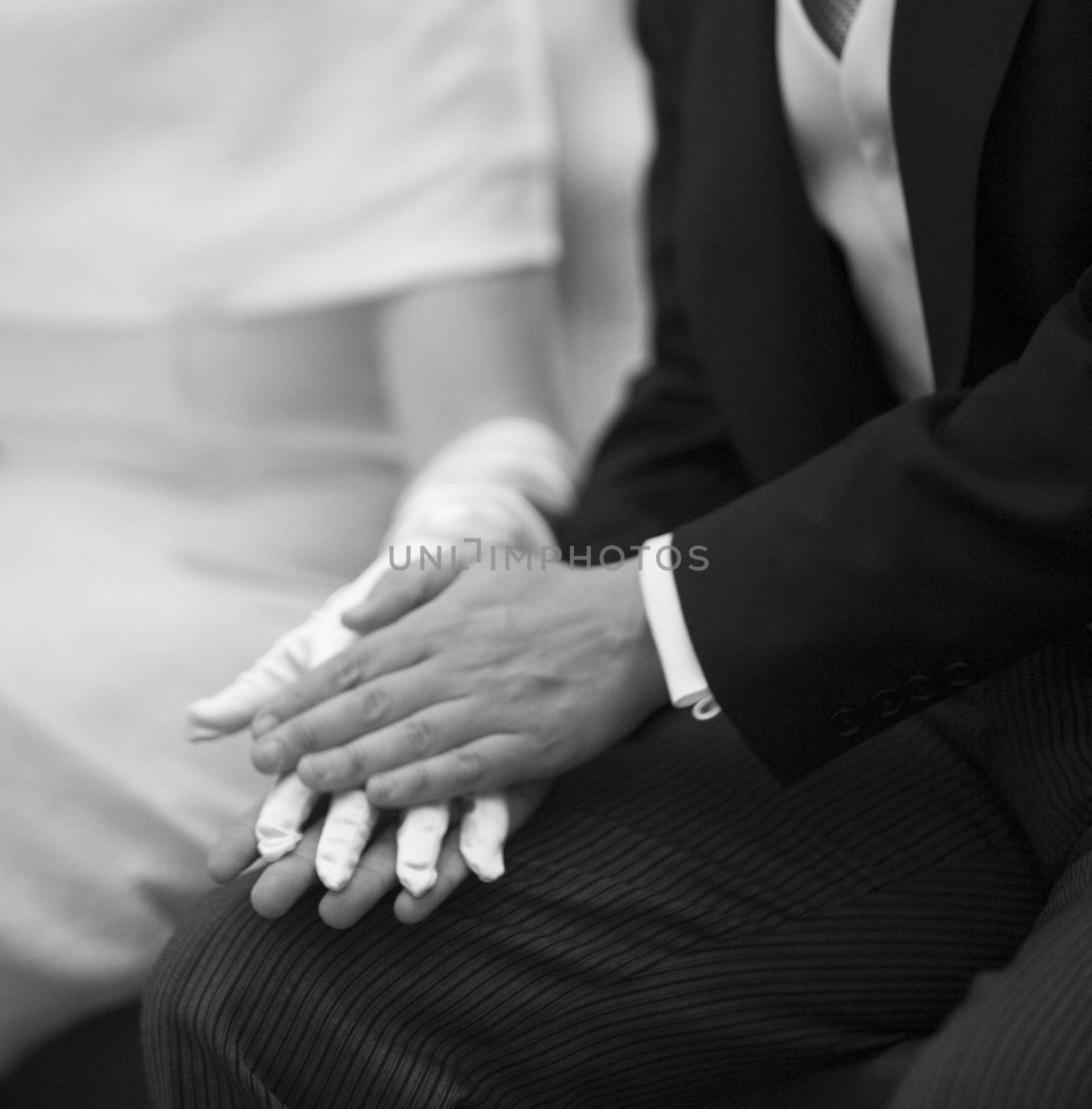 Black and white artistic digital square photo of hand of bridegroom in dark long morning suit and white shirt with cufflinks in church religious wedding marriage ceremony holding hands with the bride in white long wedding bridal dress and white gloves in Barcelona Spain. Shallow depth of with background out of focus.