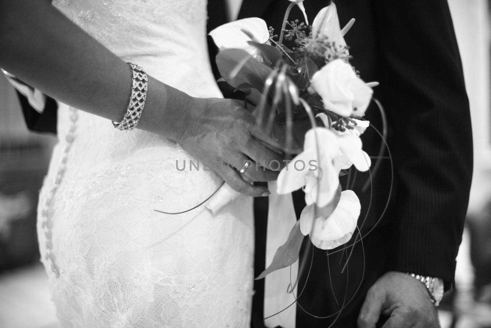 Black and white artistic digital photo of bridegroom in dark suit in wedding marriage event holding hands with the bride in white long wedding bridal dress. Shallow depth of with background out of focus. 